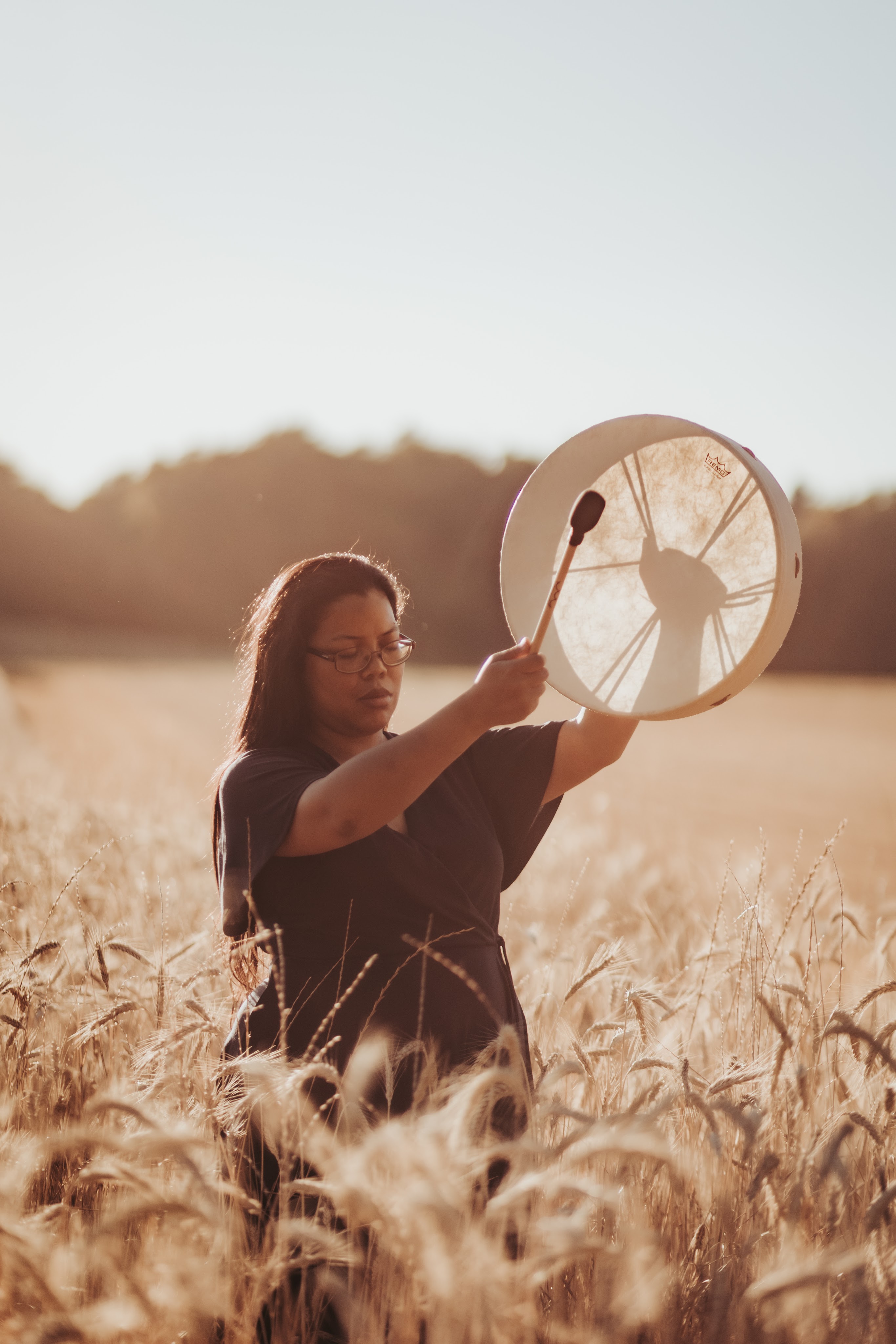 portrait d'une thérapeute en constellations familiales et systémiques tenant un tambour chamanique, vue de face, les yeux fermés, debout dans la nature en été, région Morges Renens Lausanne dans le canton de Vaud