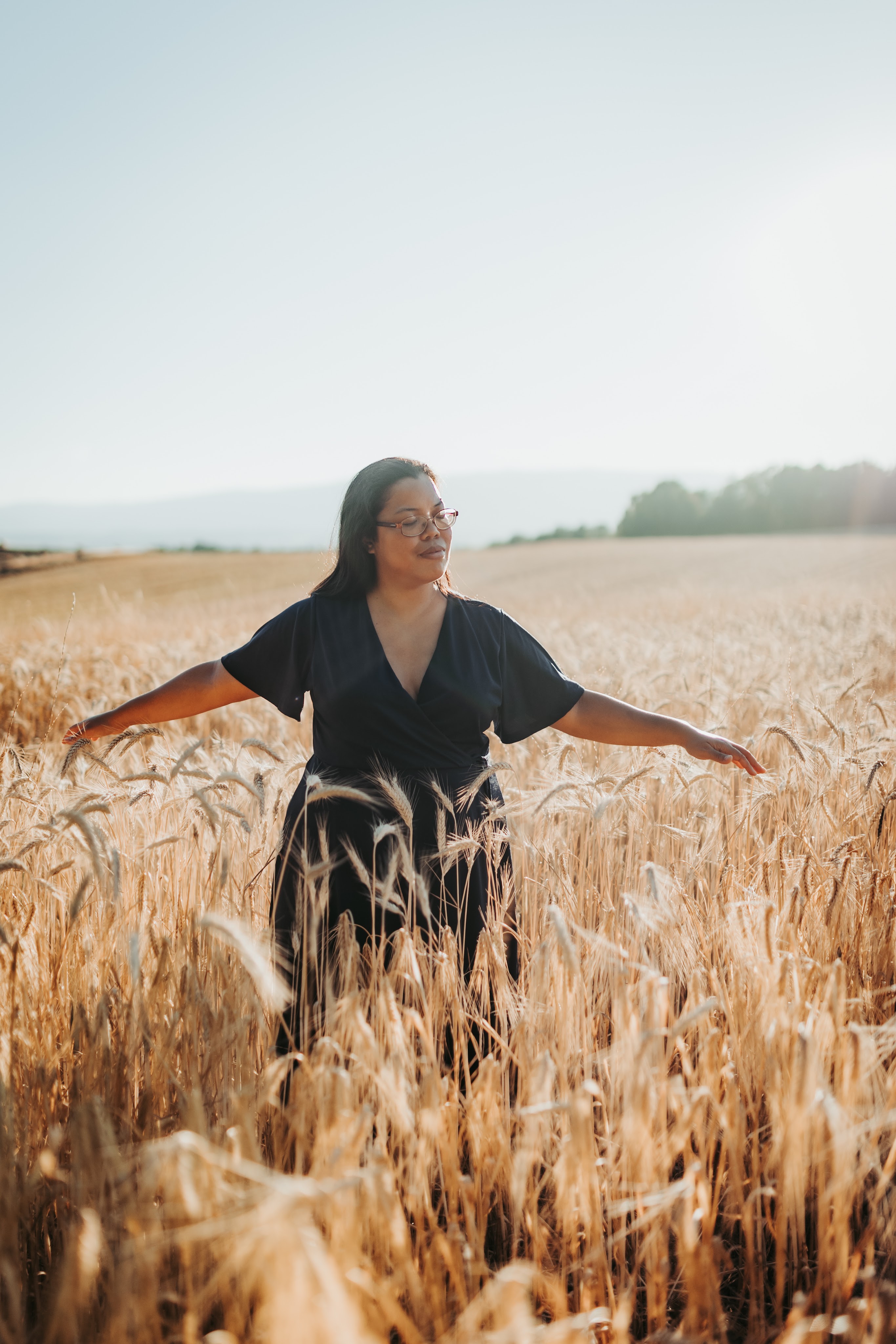 zoom sur une thérapeute en constellations familiales et systémiques et thérapie psycho-spirituelle, se tenant debout les bras écartés dans un champ situé vers Morges et Renens, près de Lausanne en Suisse, avec les yeux fermés et la forêt en arrière-plan