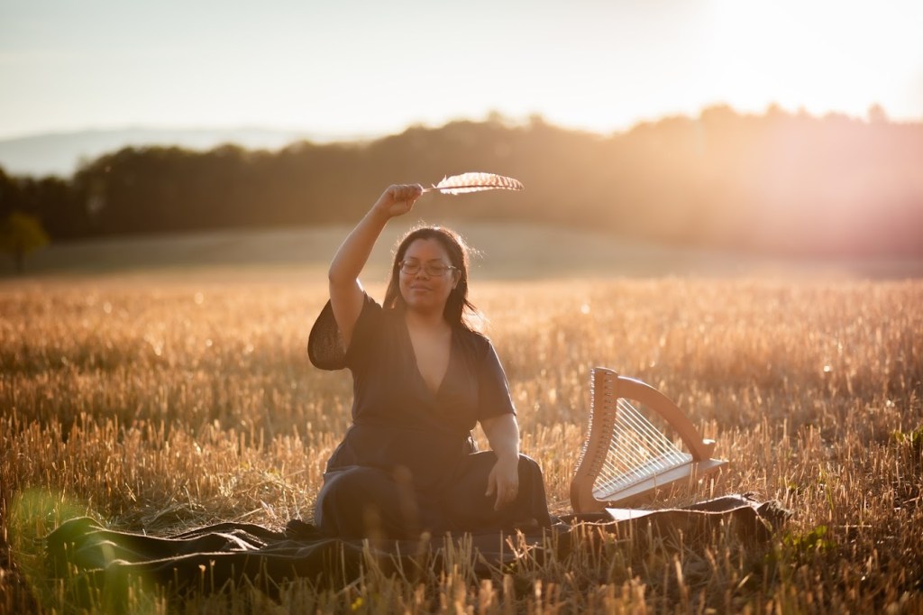 image d'une thérapeute en constellations familiales et systémiques assise dans un pré avec la forêt en arrière-plan, au soleil couchant, près d'une harpe et portant une plume, les yeux fermés, le visage doux, en train d'exercer une thérapie holistique dans le canton de Vaud, en Suisse, près de Morges, Renens et Lausanne dans le canton de Vaud, pratiquant la thérapie psycho-spirituelle