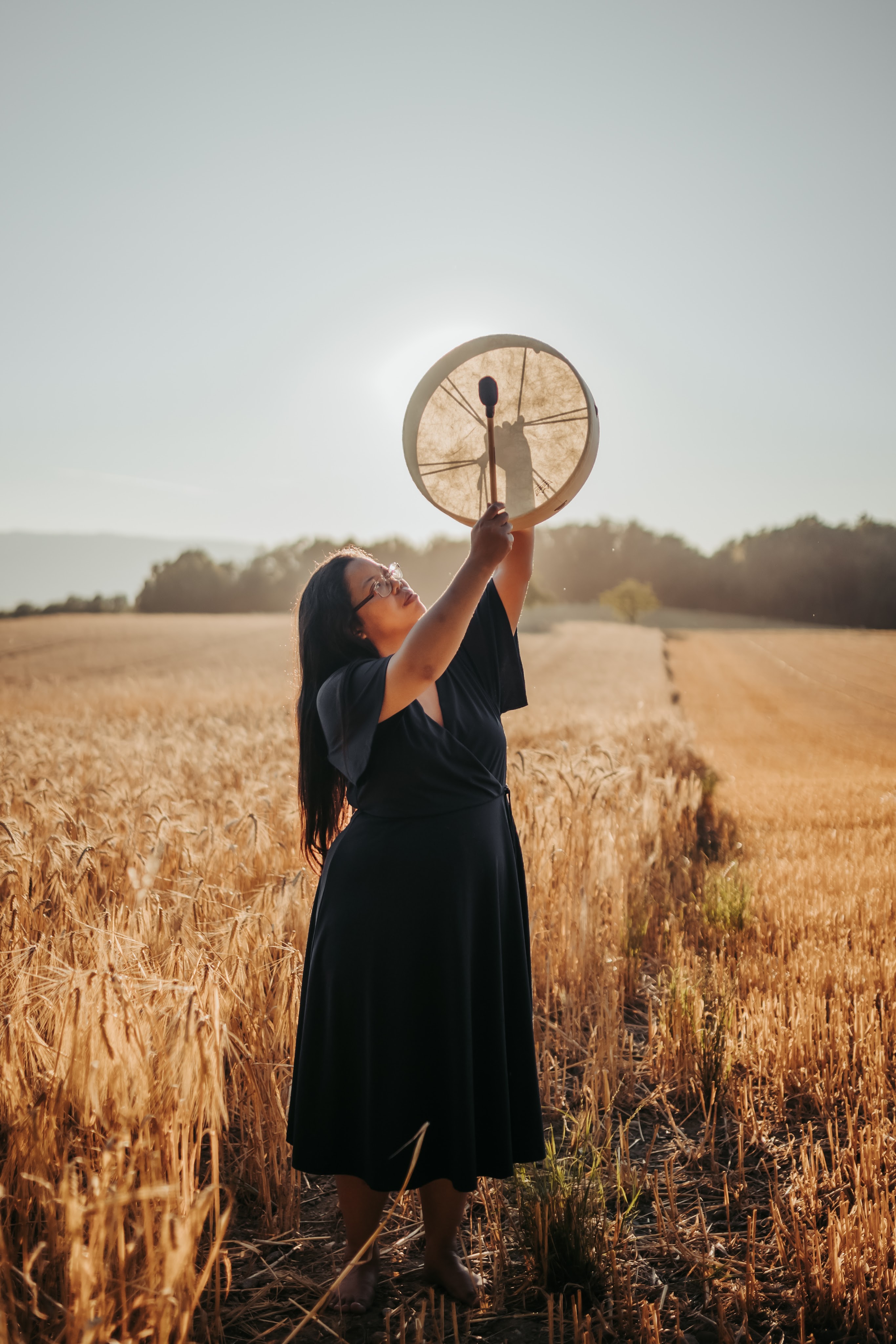thérapeute en constellations familiales et constellations systémiques se trouvant dans la nature près de Morges et Renens, dans le canton de Vaud en Suisse, à proximité de Lausanne. Debout, elle tient un tambour chamanique levé. Un champ de blé, une forêt et le soleil couchant sont visibles derrière elle.